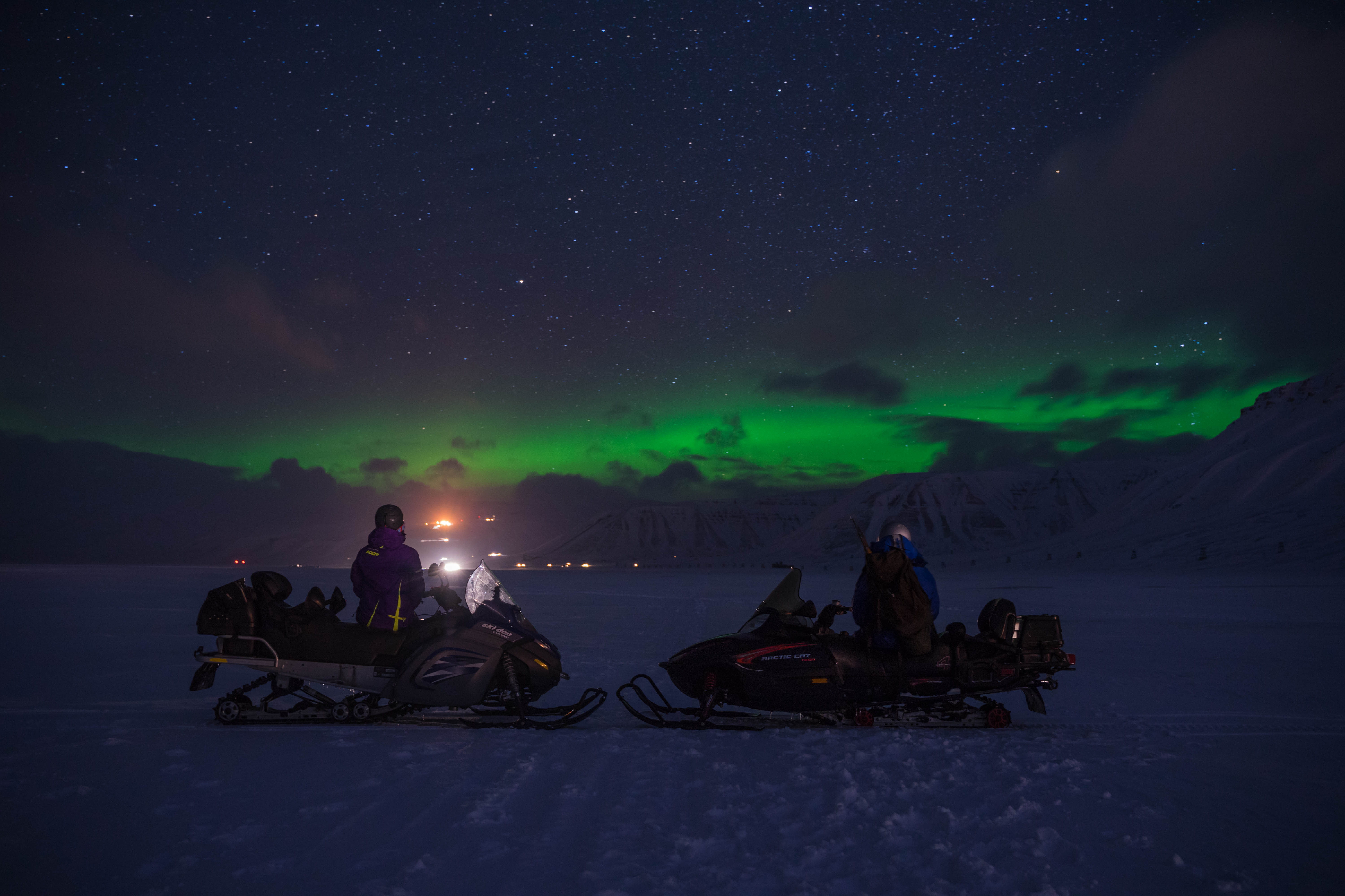 To personer på snøscootere som ser utover et landskap med nordlys på himmelen i bakgrunnen